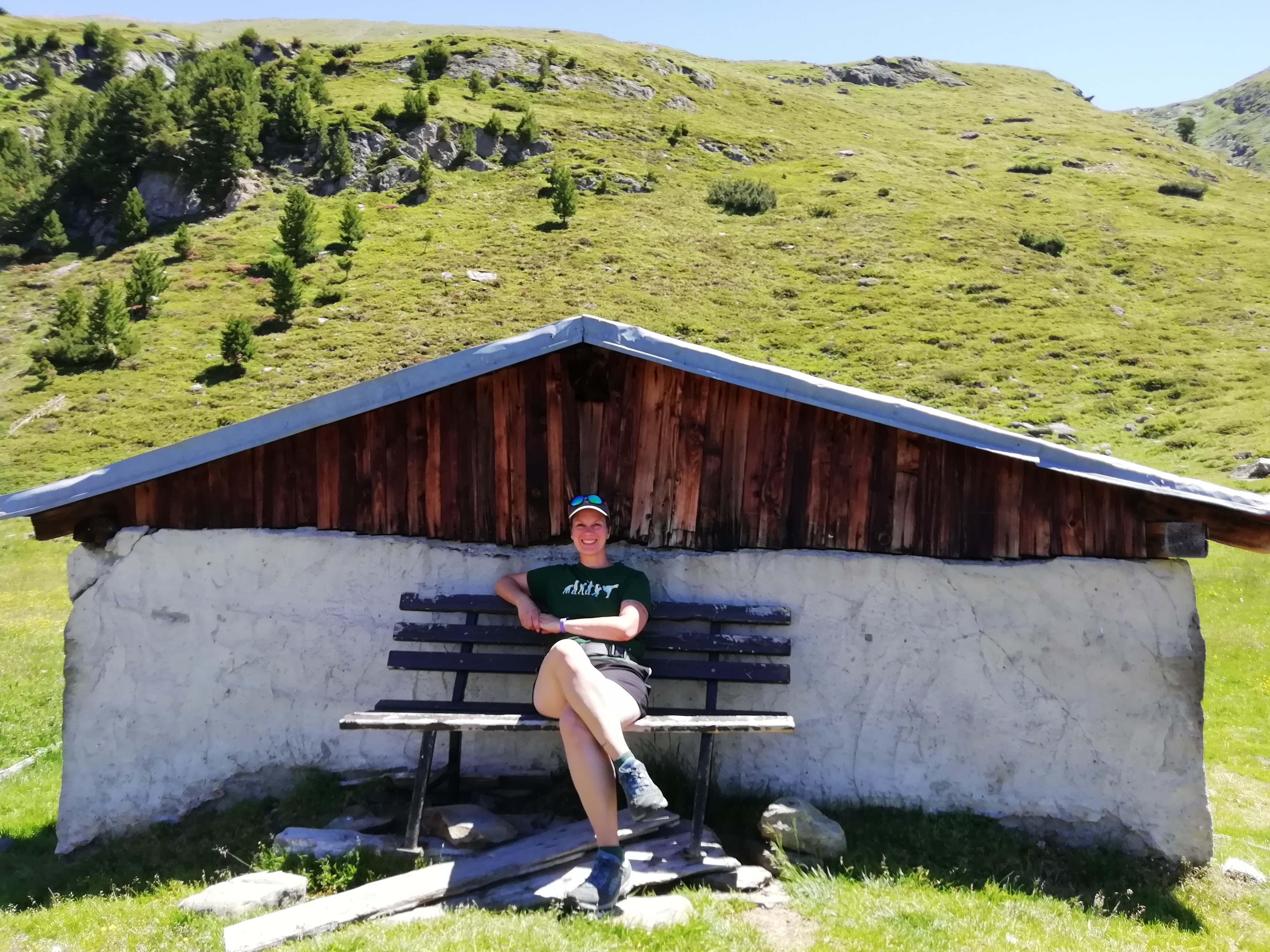 Eine niedrige, alte Hütte in den Alpen, im Hintergrund eine Bergwiese. Es ist Sommer, die Autorin sitzt lächelnd auf einer Bank davor und trägt Wanderkleidung.